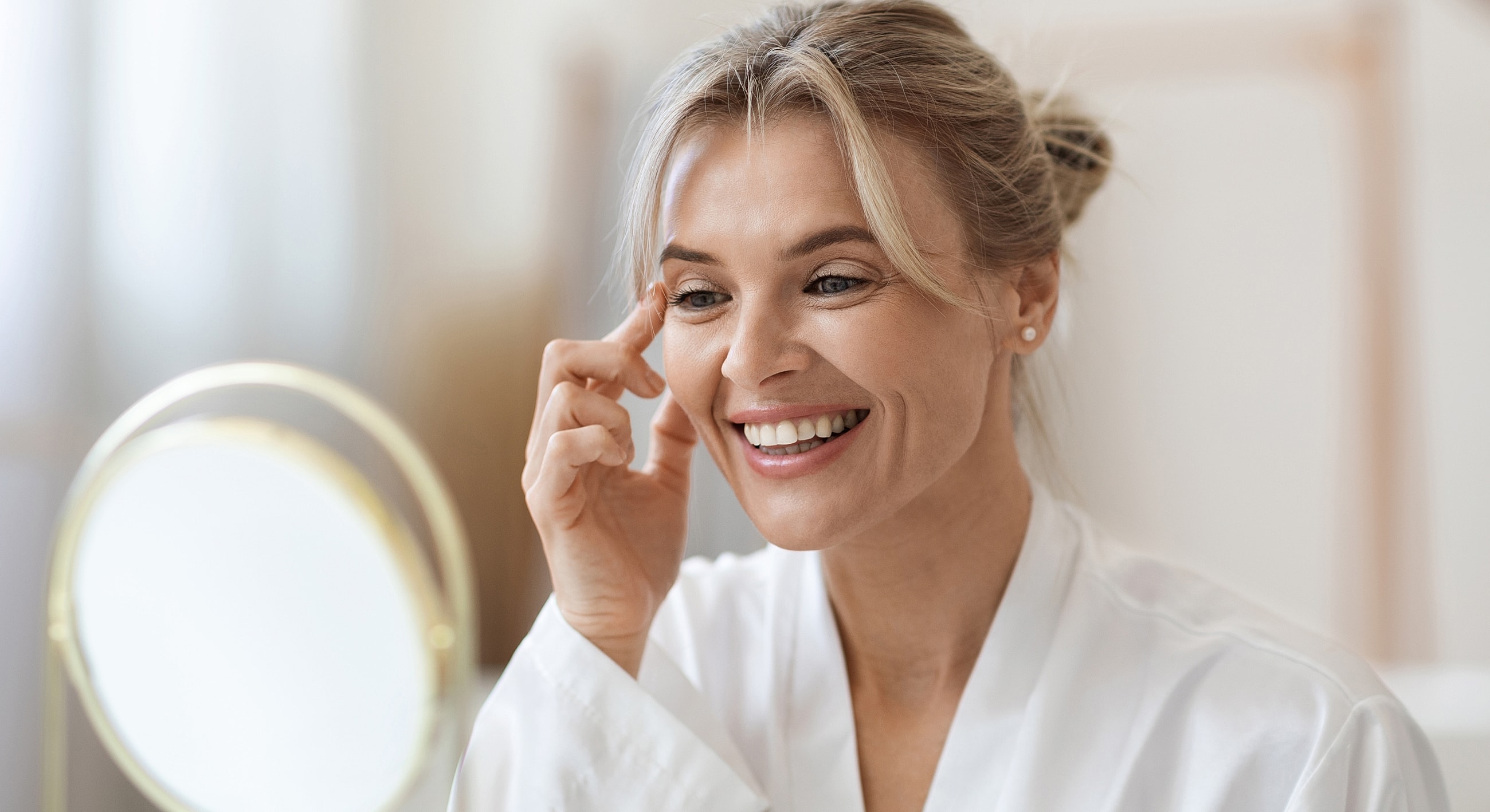 Smiling woman applying skincare in front of mirror.